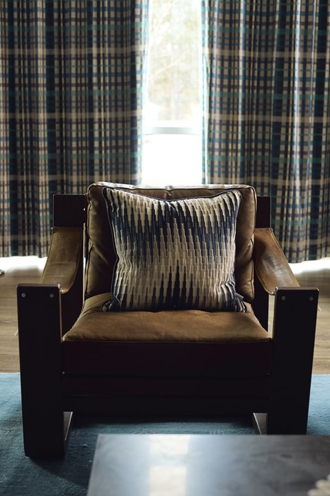 A brown chair with a striped pillow is in front of a window with blue curtains.